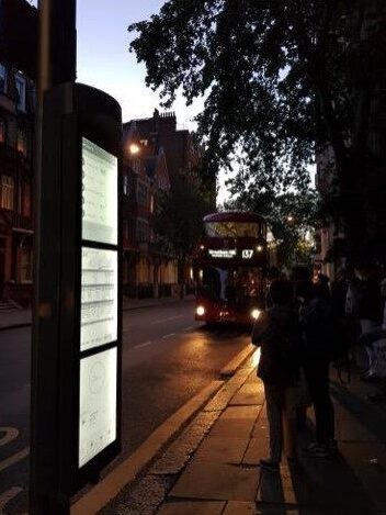 Backlit Papercast E-Paper real-time Information Timetable on a bus stop in London, England at night with double-decker bus approaching stop